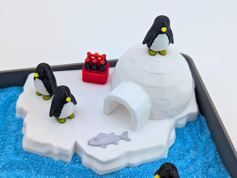 May include: A miniature arctic scene featuring a white igloo, three black and white penguins with yellow accents, and a red crate of bottles. A silver fish and blue textured base complete the diorama.