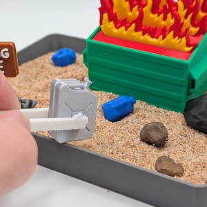 Może przedstawiać: Miniaturowa scena z zielonym śmietnikiem w ogniu, szarym metalowym kanistrem, czarną workiem na śmieci i niebieskimi plastikowymi pojemnikami. Scena jest ustawiona w tacy wypełnionej piaskiem i małymi kamieniami. Tabliczka głosi "Wszystko w porządku".