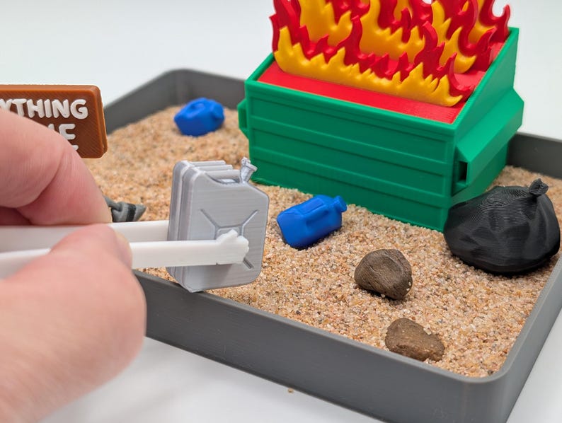 Może przedstawiać: Miniaturowa scena z zielonym śmietnikiem w ogniu, szarym metalowym kanistrem, czarną workiem na śmieci i niebieskimi plastikowymi pojemnikami. Scena jest ustawiona w tacy wypełnionej piaskiem i małymi kamieniami. Tabliczka głosi "Wszystko w porządku".