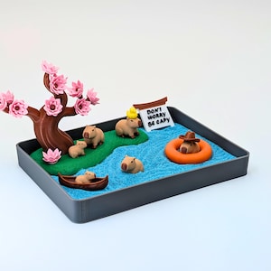 May include: A miniature diorama featuring capybaras in a serene setting. A brown tree with pink blossoms stands beside a river of blue sand. One capybara floats in an orange inner tube, and a sign reads "Don't Worry Be Capy."