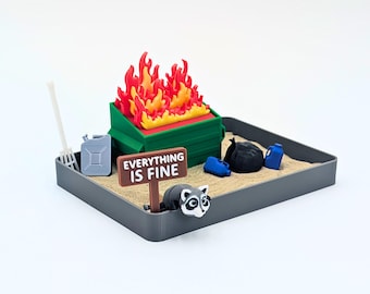 Puede incluir: Un diorama en miniatura que muestra un contenedor de basura verde envuelto en llamas rojas y amarillas brillantes. Un letrero marrón al frente dice EVERYTHING IS FINE. Una pequeña figura de mapache, un rastrillo gris, un bidón gris, jarras azules y una bolsa de basura negra.