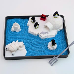 May include: A miniature arctic scene in a black tray, featuring a blue sandy sea, icebergs, and an igloo. Includes a polar bear, penguins, and a trident. The scene is a playful, decorative item.