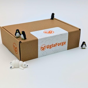 May include: A brown cardboard box with a white label featuring the text "UglaForge" and a logo. Miniature penguins and a polar bear figurine are arranged around the box. The box has a brown logo on the side.