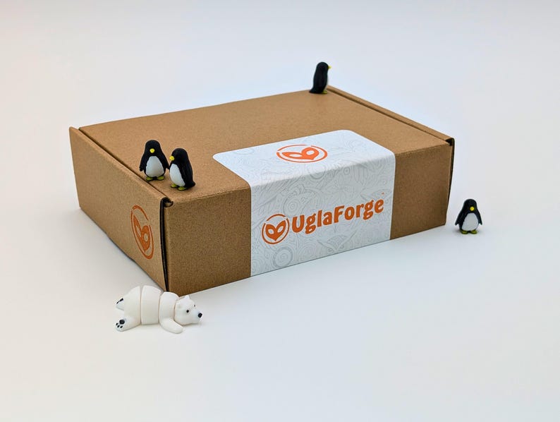 May include: A brown cardboard box with a white label featuring the text "UglaForge" and a logo. Miniature penguins and a polar bear figurine are arranged around the box. The box has a brown logo on the side.