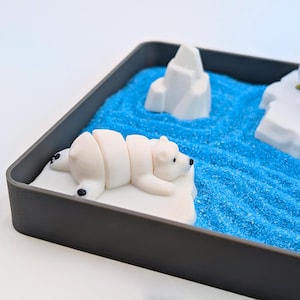 May include: A miniature arctic diorama in a gray tray. A white polar bear figurine rests on a white iceberg, with two penguin figurines on another iceberg. Blue sand represents the ocean, and a white iceberg is in the background.