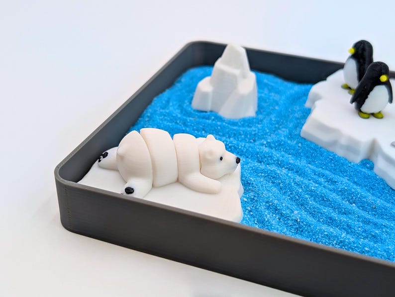 May include: A miniature arctic diorama in a gray tray. A white polar bear figurine rests on a white iceberg, with two penguin figurines on another iceberg. Blue sand represents the ocean, and a white iceberg is in the background.