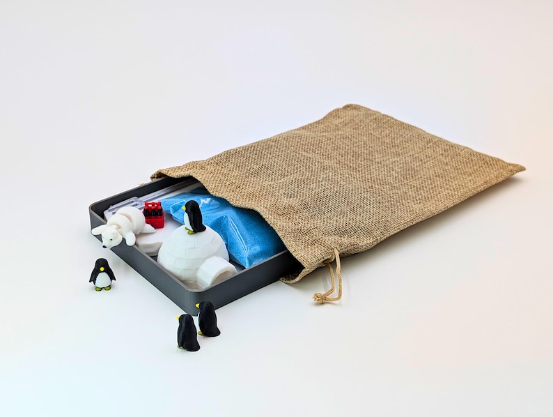 May include: A gray metal box containing a miniature polar scene with a white igloo, polar bear, and penguins. A burlap bag is partially open, revealing the contents. The scene includes red and blue accents.