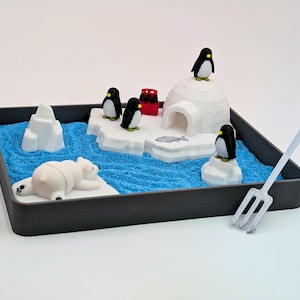 May include: A miniature arctic scene in a dark gray tray. It features a polar bear, penguins, an igloo, and icebergs on a blue, sandy surface. A silver rake is positioned to the right. The scene is a playful representation of the arctic environment.
