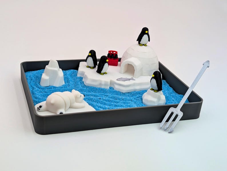 May include: A miniature arctic scene in a dark gray tray. It features a polar bear, penguins, an igloo, and icebergs on a blue, sandy surface. A silver rake is positioned to the right. The scene is a playful representation of the arctic environment.