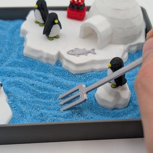 May include: A miniature diorama scene featuring a polar bear, penguins, and an igloo on a blue sandy surface. The scene includes a small red container and a fish. A hand holds a silver fork over the scene.