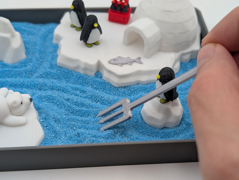 May include: A miniature diorama scene featuring a polar bear, penguins, and an igloo on a blue sandy surface. The scene includes a small red container and a fish. A hand holds a silver fork over the scene.