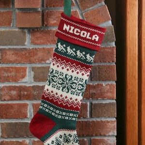 Puede incluir: Un calcetín de Navidad de punto rojo y verde con un patrón de copo de nieve blanco. El calcetín tiene el nombre "Nicola" bordado en el puño.