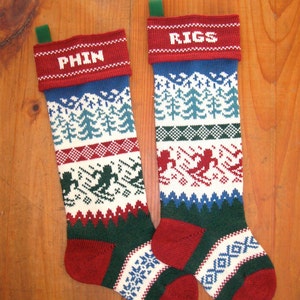 Op de afbeelding: Twee gebreide kerstsokken met de namen "PHIN" en "RIGS" in witte letters. De sokken hebben een rode manchet, blauwe en witte boompatronen en skiërs. De onderkant is rood en groen met een wit en blauw geometrisch ontwerp. Elke sok heeft een groene lus.