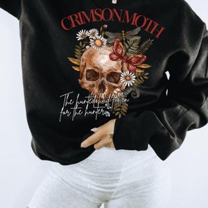 Puede incluir: Sudadera negra con un diseño de calavera, margaritas y una mariposa. El texto "CRIMSON MOTH" está encima de la calavera, y la frase "The hunted had fallen for the hunter" está debajo. El diseño utiliza tonos rojos, marrones y blancos.