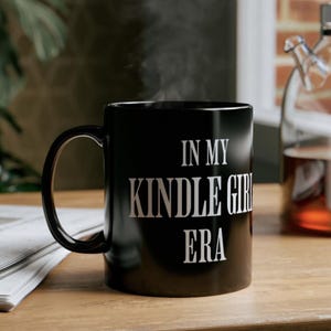 Puede incluir: Taza de cer&aacute;mica negra con el texto blanco "IN MY KINDLE GIRL ERA". La taza humea, lo que sugiere una bebida caliente. La taza est&aacute; sobre una superficie de madera, con un peri&oacute;dico y una botella de vidrio en el fondo.
