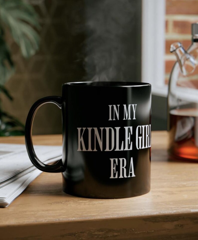 Puede incluir: Taza de cer&aacute;mica negra con el texto blanco "IN MY KINDLE GIRL ERA". La taza humea, lo que sugiere una bebida caliente. La taza est&aacute; sobre una superficie de madera, con un peri&oacute;dico y una botella de vidrio en el fondo.