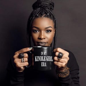 Puede incluir: Una taza de caf&eacute; negra con el texto "IN MY KINDLE GIRL ERA" en blanco. La taza la sostiene una persona de piel oscura, que lleva una camiseta negra y anillos. El fondo es de color gris oscuro.