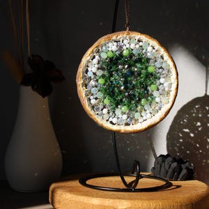 May include: A round, green, white and brown beaded mandala hanging from a black metal stand. The mandala is made of various sizes and shapes of beads, including green, white and clear beads.