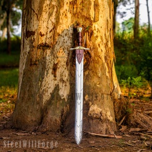 May include: A silver-colored metal sword with a dark brown handle and a gold-toned crossguard. The blade features a central ridge and decorative etched lines. A brown leather sheath with a dark trident emblem is attached to the upper blade.