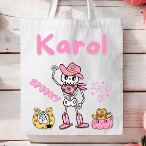 Puede incluir: Bolsa de lona blanca con un esqueleto rosa que lleva un sombrero de vaquero rosa y botas. La bolsa tiene el nombre "Karol" escrito en letras rosas y la palabra "Spooky" escrita en rosa. La bolsa también tiene una herradura amarilla con una telaraña y una calabaza rosa con un sombrero de vaquero.