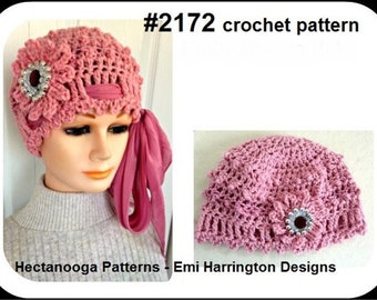 May include: A pink crochet hat with a floral design and a decorative jewel. The hat has a matching pink ribbon and is displayed on a mannequin head. The image also includes the text "#2172 crochet pattern".