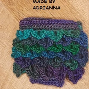May include: Handmade crochet item in shades of blue, green, and purple. The item has a scalloped edge and a textured, layered design. The text "CUSTOMER SUBMITTED PHOTO" and "MADE BY ADRIANNA" are visible.
