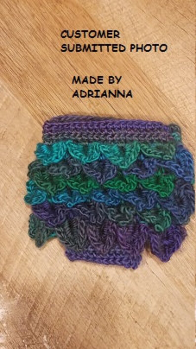 May include: Handmade crochet item in shades of blue, green, and purple. The item has a scalloped edge and a textured, layered design. The text "CUSTOMER SUBMITTED PHOTO" and "MADE BY ADRIANNA" are visible.