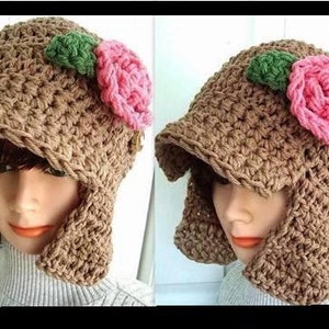 May include: A brown crocheted hat with a wide brim and ear flaps. The hat is decorated with two pink and green fabric flowers.