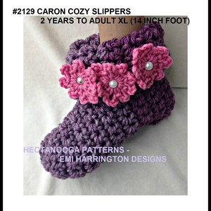 May include: A handmade purple crochet slipper with pink flower accents and pearl centers. The slipper is designed for 2 years to adult XL (14 inch foot). The text on the image reads "#2129 CARON COZY SLIPPERS" and "HECTANOOGA PATTERNS - EMI HARRINGTON DESIGNS."
