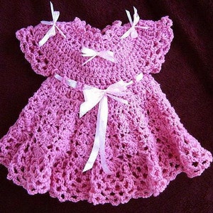 May include: A pink crocheted baby dress with white satin bows. The dress has a scalloped hem and a delicate crocheted pattern.