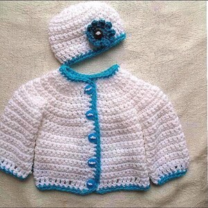 May include: White crocheted baby cardigan with blue trim and buttons. Matching white crocheted hat with a blue flower.