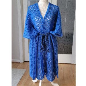 Pode incluir: Uma bata de croch&ecirc; azul com cinto de la&ccedil;o. A bata est&aacute; em um manequim branco e o texto "MADE BY TRISH BRESOLIN" est&aacute; na parte superior da imagem.