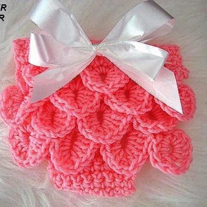 May include: A handmade, coral-colored crochet diaper cover with a scale-like pattern. The cover is adorned with a large, white satin bow. The text "DIAPER COVER" is visible in the upper left corner.