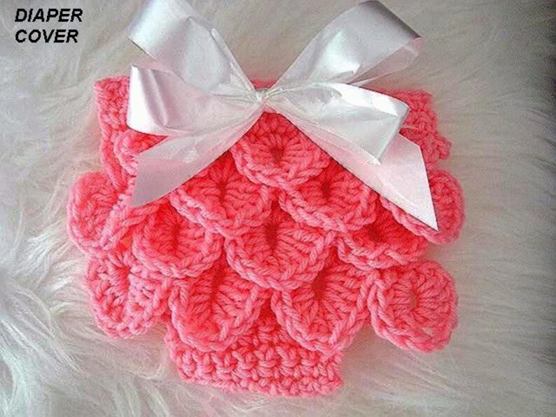 May include: A handmade, coral-colored crochet diaper cover with a scale-like pattern. The cover is adorned with a large, white satin bow. The text "DIAPER COVER" is visible in the upper left corner.