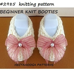 Könnte beinhalten: Paar cremefarbene gestrickte Baby-Booties mit rosa Schleifen und Perlenakzenten. Der Text "#2985 knitting pattern BEGINNER KNIT BOOTIES" ist sichtbar. Die Booties werden auf weißen Formen präsentiert.