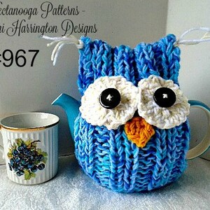 Pode incluir: Capa de chaleira de coruja de malha azul com olhos de crochê branco e bico amarelo. A capa está em uma chaleira azul com uma caneca branca ao lado. O texto "#967" está no canto superior esquerdo da imagem.