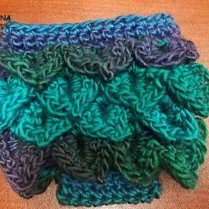 May include: Handmade crochet item in shades of teal, green, and blue, resembling dragon scales. The item is crafted with textured yarn, creating a three-dimensional effect. The text "MADE BY ADRIANNA" is visible in the image.