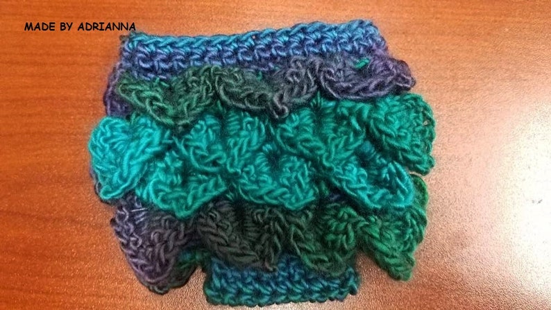 May include: Handmade crochet item in shades of teal, green, and blue, resembling dragon scales. The item is crafted with textured yarn, creating a three-dimensional effect. The text "MADE BY ADRIANNA" is visible in the image.