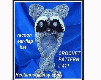 May include: A crocheted raccoon ear-flap hat in shades of gray, black, and white. The hat features a raccoon face design with ear flaps and the text "raccoon ear-flap hat". The text "CROCHET PATTERN #411" is also visible.