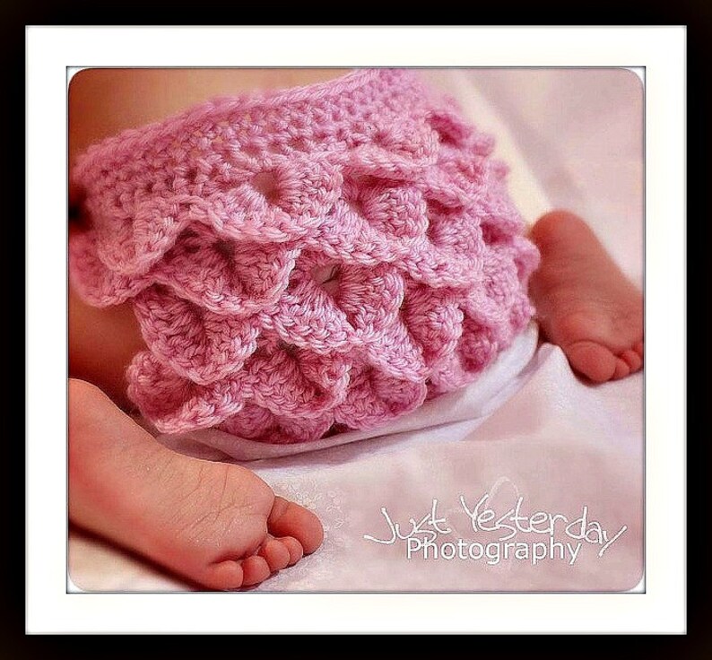 May include: A close-up of a pink crocheted diaper cover with a scalloped design. The cover is on a baby, with the baby's feet visible. The image is set against a soft, white background.