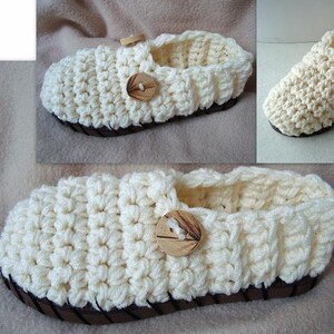 Puede incluir: Un par de zapatillas de crochet de color crema con un botón de madera en cada una. Las zapatillas tienen un ribete marrón alrededor del borde.