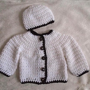 May include: White crocheted baby cardigan with black trim and buttons. The cardigan is paired with a matching white crocheted hat with black trim.