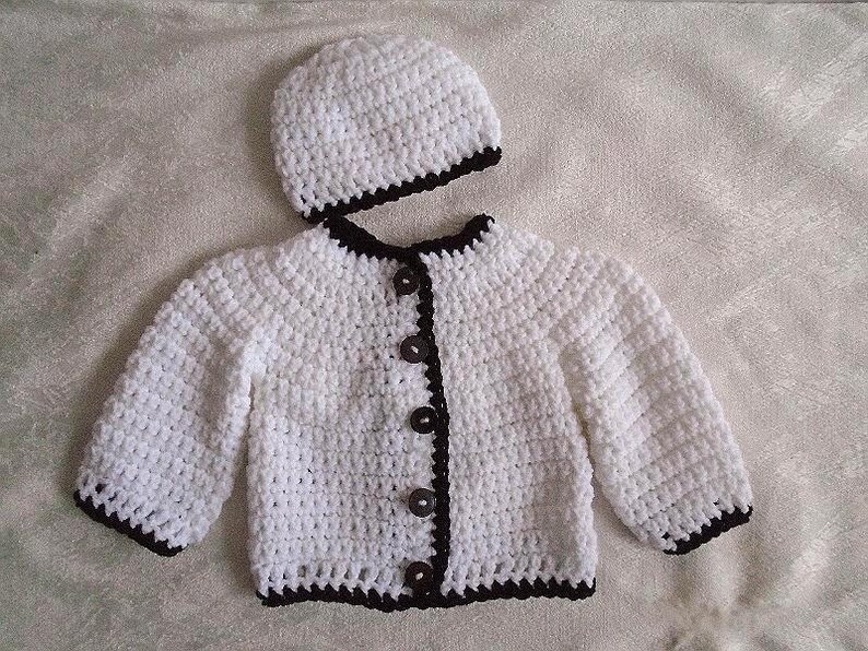 May include: White crocheted baby cardigan with black trim and buttons. The cardigan is paired with a matching white crocheted hat with black trim.