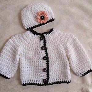May include: White crocheted baby cardigan with black trim and buttons. Matching white crocheted hat with a pink flower.