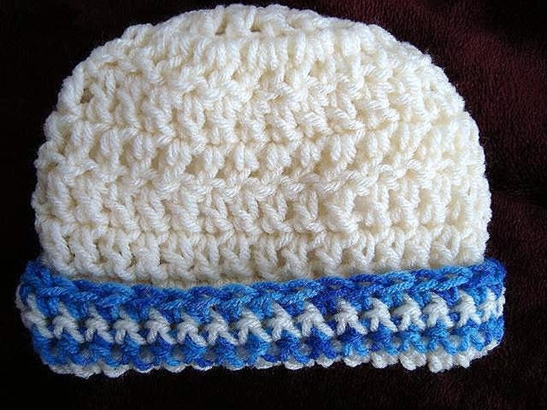 May include: A crocheted beanie hat. The main body of the hat is a creamy white color, with a textured stitch pattern. The brim is a contrasting blue and white, adding a pop of color. The hat is handmade and suitable for cold weather.