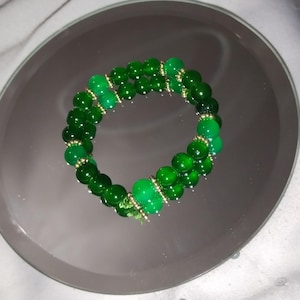 May include: A green beaded bracelet with gold accents. The bracelet is made of round beads and is wrapped twice around the wrist.