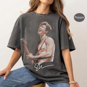 Könnte beinhalten: Pfefferfarbenes Grafik-T-Shirt mit einem Gitarre spielenden und lächelnden Musiker. Das Bild ist in Sepia-Tönen gehalten, mit der Unterschrift des Musikers in weißer Schrift darunter. Das Shirt hat kurze Ärmel und eine lockere Passform.