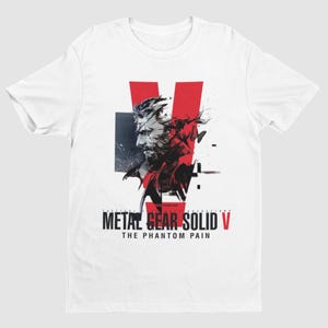 Puede incluir: Camiseta blanca con un gráfico rojo y negro de un personaje del videojuego Metal Gear Solid V: The Phantom Pain. El gráfico presenta la cara del personaje y una "V" roja en el fondo.