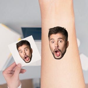 May include: A temporary tattoo of a man's face with an open mouth and surprised expression is applied to an arm. A small card with the same image is held nearby. The tattoo is a realistic depiction of a man with dark hair and a beard.