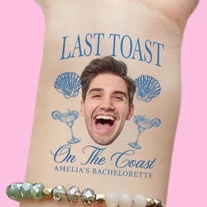 May include: A temporary tattoo with the text "LAST TOAST" above a man's face, two seashell illustrations, and two margarita glasses. Below, the text reads "On The Coast" and "AMELIA'S BACHELORETTE". The tattoo is on a light skin tone.